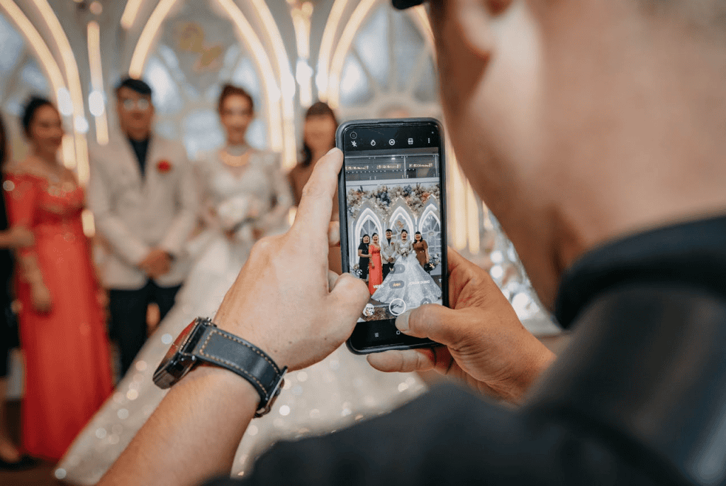A person uses a smartphone to capture four people, including a bride in a white dress, posing at a decorated wedding venue with arched lights—perfect for instant sharing on your favorite media-sharing app.