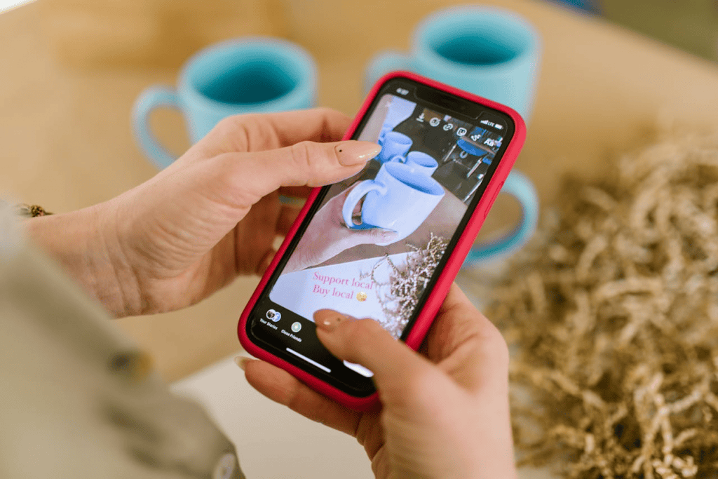 A person holding a smartphone with a red case uses an AI-powered media-sharing app to snap a photo of blue mugs on a table. The screen displays the mugs, the words Support local, buy local, and straw decor in the background.