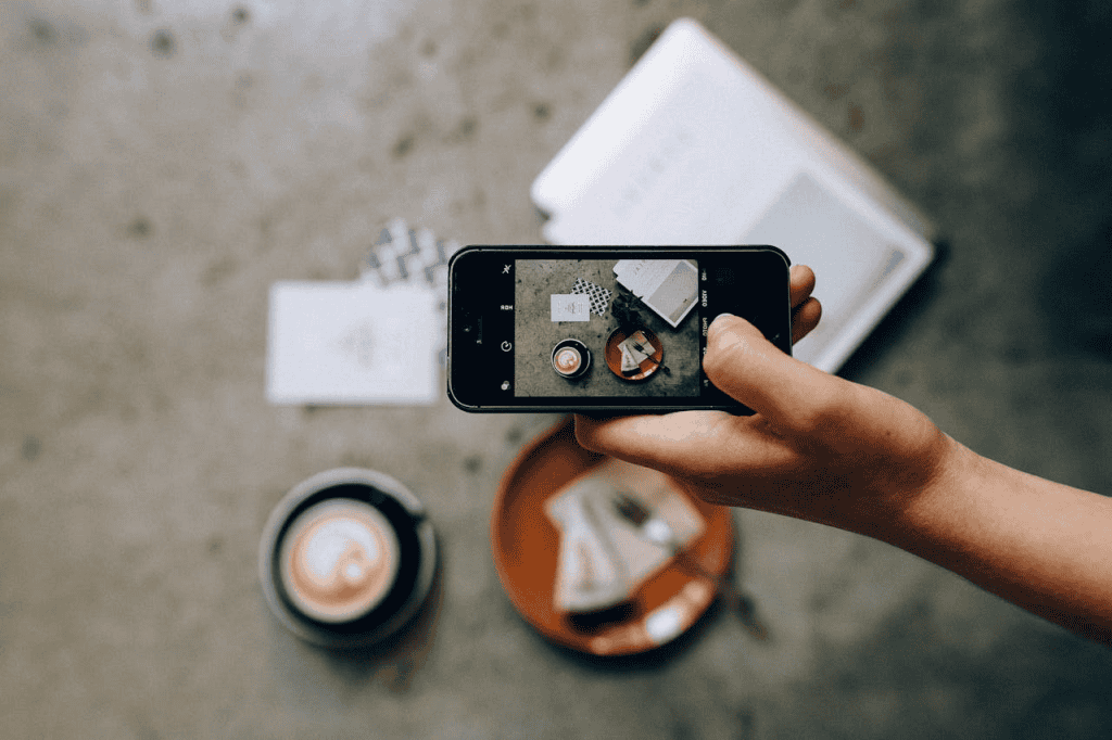 A person holds a smartphone, taking a photo of a table with coffee and dessert on a plate, seen from above—perfect for sharing on an AI-powered media-sharing app that enhances functionality and creativity.