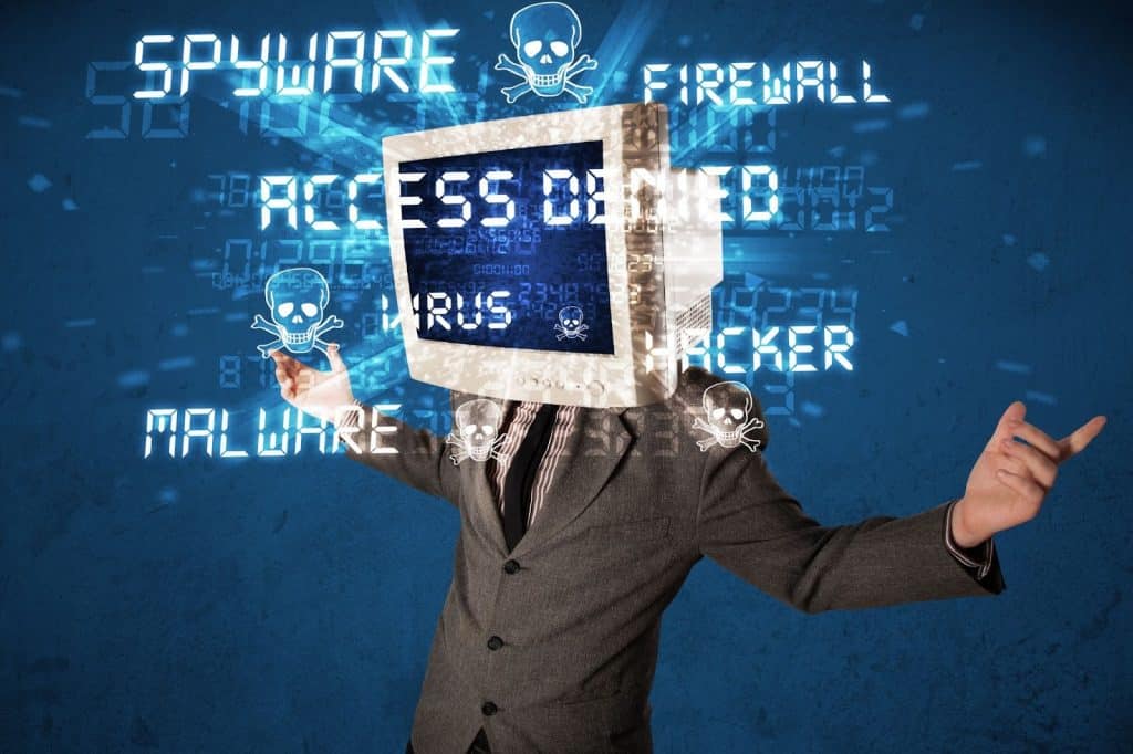 A person in a suit with a computer monitor for a head stands with arms open. The screen displays Access Denied, surrounded by digital graphics and words like Spyware, Virus, Hacker, and Malware on a blue background.