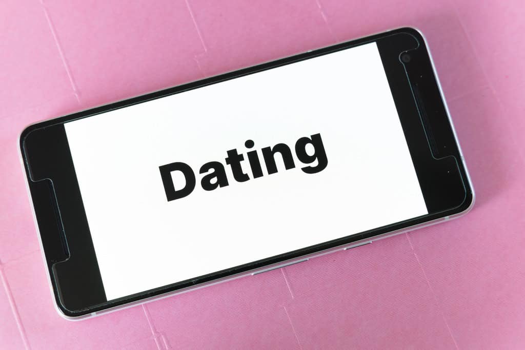 A smartphone lying on a pink surface displays the word Dating in bold black letters on its screen.