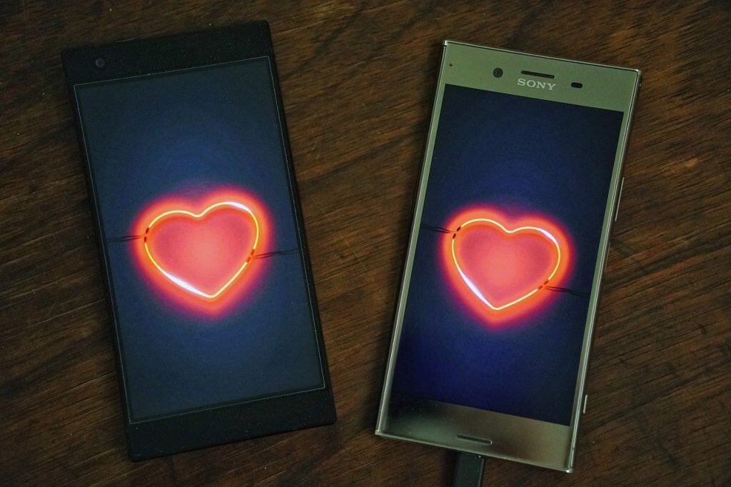 Two smartphones lying on a wooden surface, each displaying a glowing neon heart on their screens, symbolizing a digital or long-distance romantic connection.