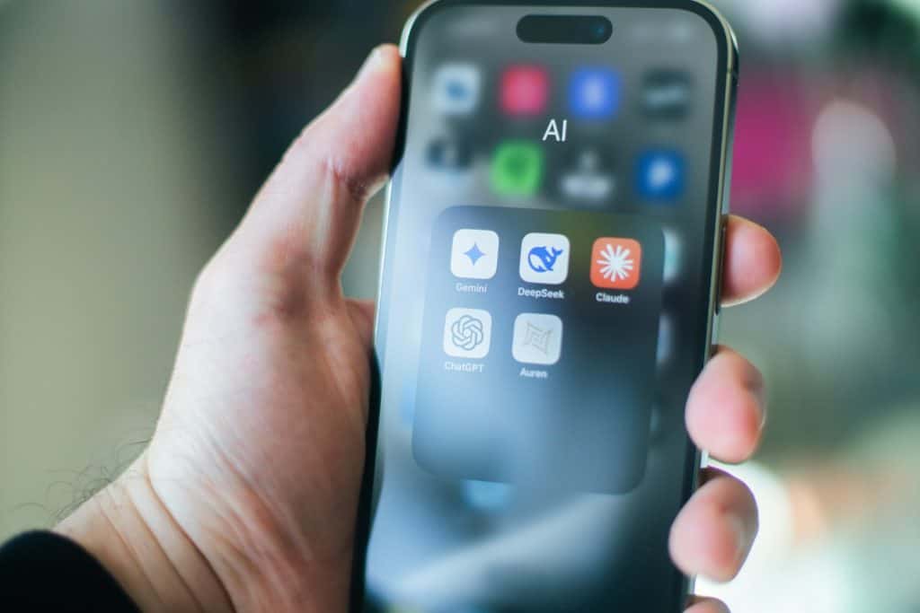 A hand holding a smartphone displaying a folder labeled AI with six app icons: Gemini, DeepSeek, Claude, ChatGPT, and two others, on the screen.